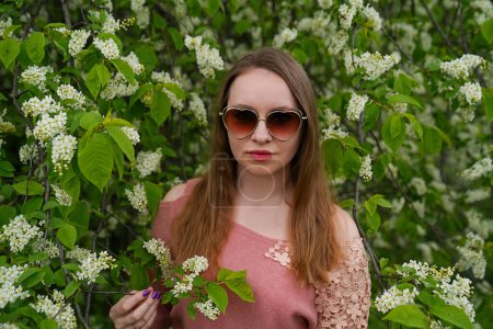 beautiful blond natural hair woman wears sunglasses in pink outfit is posing in botanical garden park near blooming bird cherry mayday tree with flowers. Spring and purity, natural beauty