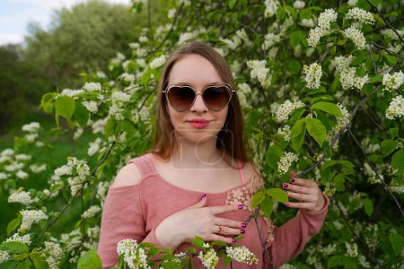 beautiful blond natural hair woman wears sunglasses in pink outfit is posing in botanical garden park near blooming bird cherry mayday tree with flowers. Spring and purity, natural beauty
