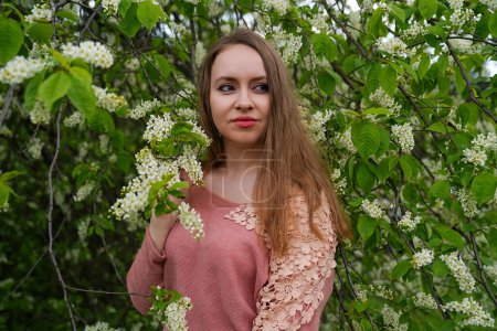 beautiful blond natural hair woman in pink outfit is posing in botanical garden park near blooming bird cherry mayday tree with flowers. Spring and purity, natural beauty