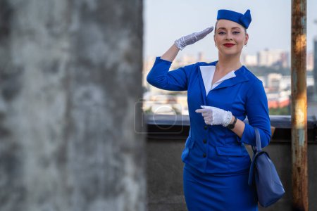 Stewardess in blauer Vintage-Uniform mit Mütze, Handschuhen, Handtasche macht einen Gruß. Stewardess berührt ihre Hand am Hut. Arbeit in der Luftfahrt. Kundendienstbranche, Reisen mit dem Flugzeug um die Welt