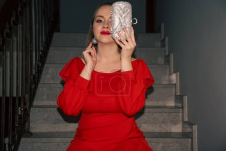 glamour jeune belle femme en robe rouge, avec sac en argent et boucles d'oreilles en argent est assis sur l'escalier, style rétro vintage
