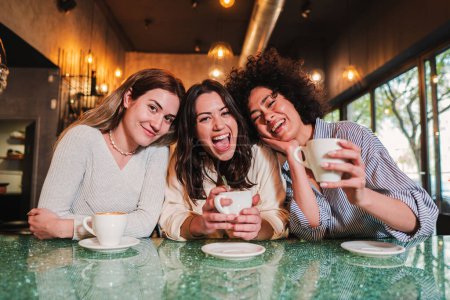 Tres mujeres jóvenes sonriendo mirando la cámara en una cafetería. Un grupo de chicas felices multirraciales divirtiéndose juntas tomando un descanso en un restaurante. Foto de alta calidad