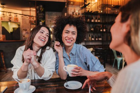 Téléchargez les photos : A group of young women talking and laughing sitting on a coffee shop or restaurant. Two ladies smiling and looking his friend holding a cup. Three girls having fun cahtting on a bar. Lifestyle concept - en image libre de droit