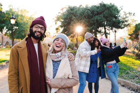 Foto de Young lovely couple smiling and laughing looking at camera, at background three friends taking a selfie photo with a cellphone, wearing scarf, hats, coats and winter or autumn clothes. Lifestyle - Imagen libre de derechos