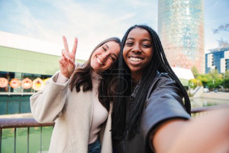 Foto de A couple of young multiracial women having fun taking a selfie photo portrait. Front view of one happy african american girl and her friend, a pretty caucasian brunette smiling doing the peace sign - Imagen libre de derechos