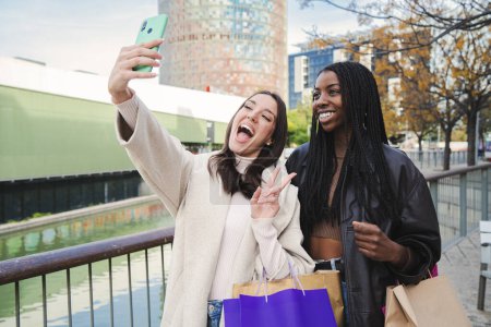 Photo for Two multiethnic young women smiling and having fun taking a selfie portrait with a smart phone after shopping and sharing in the social media. A couple of carefree girls doing a photo with a cellphone - Royalty Free Image
