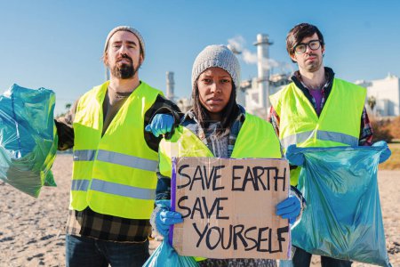 Foto de Equipo de activistas o voluntarios, recogiendo basura de la playa, sosteniendo un cartel que dice salvar la tierra sálvate a ti mismo. Un grupo de jóvenes limpia la basura de la arena. Foto de alta calidad - Imagen libre de derechos