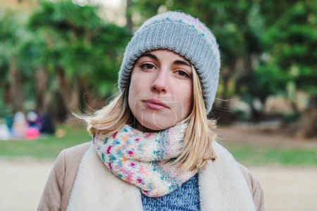 Foto de Front view of young serious blonde girl with a beanie hat looking pensive at camera standing in a park outdoors. Close up portrait of caucasian woman with autumnal clothes. High quality photo - Imagen libre de derechos