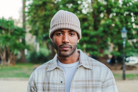Foto de Close up portrait of young attractive african american man with beanie hat looking serious at camera. Front view of a hispanic guy standing in a park outdoors with sad attitude. High quality photo - Imagen libre de derechos