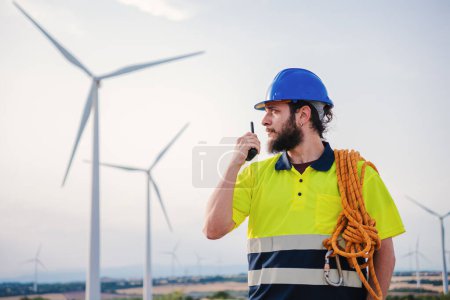 Téléchargez les photos : Renewable energy engineer or windmill turbine technician working and talking by walkie talkie, wearing a helmet and professional security clothes. Wind energy concept. High quality photo - en image libre de droit