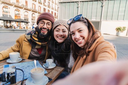 Téléchargez les photos : Group of young tourist people smiling and having fun taking a selfie portrait looking at camera. Front view of multiracial friends doing a photo in a coffee shop or restaurant terrace. Lifestyle - en image libre de droit