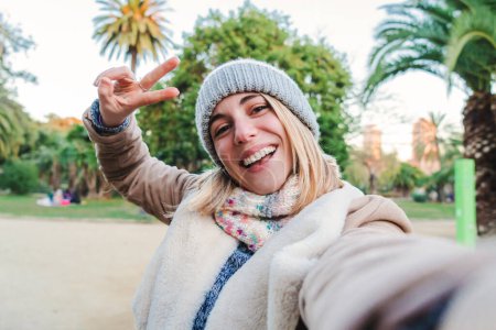 Foto de One happy caucasian blonde girl smiling taking a selfie portrait with a cellphone. European happy young woman doing the peace sign with the fingers standing in a park outdoors. Lifestyle concept. High - Imagen libre de derechos