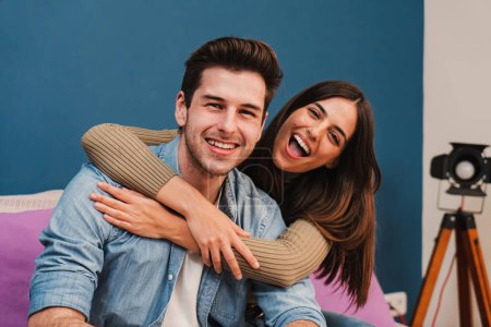 Foto de Close up portrait of happy caucasian young couple hugging and smiling together in a couch at home. Beautiful brunette wife embracing her husband resting in a sofa indoors. High quality photo - Imagen libre de derechos
