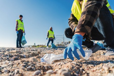 Téléchargez les photos : Volontaire mans main avec des gants ramasser les déchets en plastique de la plage pour nettoyer le sable avec ses collègues militants Recyclage concept. Photo de haute qualité - en image libre de droit