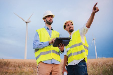Téléchargez les photos : Deux ingénieurs d'éoliennes travaillant ensemble, un technicien utilisant un dispositif tablette pour vérifier l'efficacité de son contremaître pointant quelle génératrice doit être réparée. Concept d'énergie renouvelable. Haut - en image libre de droit