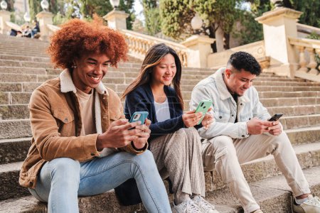Téléchargez les photos : Groupe multiracial jeunes amis appréciant et souriant en utilisant leur application de téléphone mobile assis dans les escaliers à l'extérieur. Trois heureux adolescents multiethniques s'amusent à jouer à des jeux avec leur appareil de téléphone portable - en image libre de droit