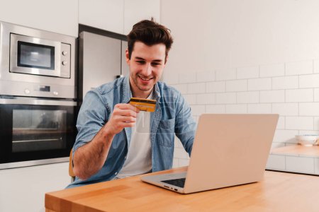 Photo for Handsome caucasian young male smiling and holding a credit card to pay for a online shopping purchase using a laptop computer at home kitchen. Banking finance concept. High quality photo - Royalty Free Image