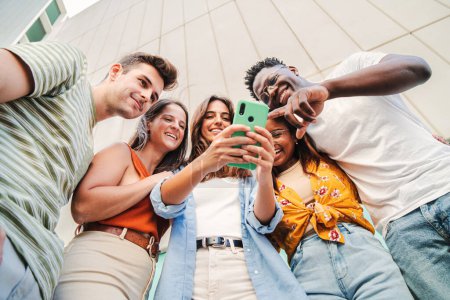 Téléchargez les photos : Vue en angle bas du groupe multiracial de jeunes amis appréciant et souriant à l'aide de leur application de téléphone mobile sur le campus universitaire. Divers adolescents s'amusent à regarder du contenu sur les médias sociaux en plein air - en image libre de droit