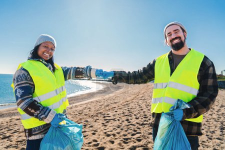 Foto de Dos voluntarios multirraciales golpeando puños después de recoger basura y suciedad de plástico de la arena en la playa. Un par de activistas ecologistas sonriendo de pie mirando a la cámara. Concepto ecológico - Imagen libre de derechos