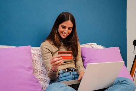 Téléchargez les photos : Jeune femme brune caucasienne réservant une chambre à l'aide d'un ordinateur portable et payant par carte de crédit assis sur un canapé à la maison à l'intérieur. Une femme achetant en ligne sur Internet. Concept de finance. Haut - en image libre de droit