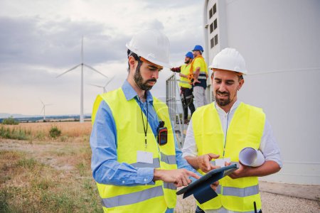 Foto de Grupo de ingenieros profesionales de molinos de viento contratistas que trabajan y utilizan una tableta para ver el proyecto de la construcción de la hélice del generador de energía renovable con casco y chaleco de seguridad - Imagen libre de derechos