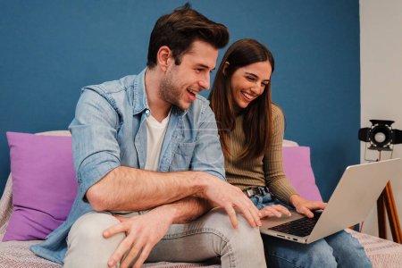 Téléchargez les photos : Caucasien jeune couple naviguant sur Internet à l'aide d'un ordinateur portable assis sur un canapé dans leur maison. Des amis heureux souriant et s'amusant à regarder des vidéos sur les médias sociaux dans leur appartement à l'intérieur - en image libre de droit