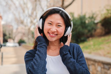 Téléchargez les photos : Une jeune femme chinoise écoutant de la musique avec un casque utilisant une connexion sans fil. Heureuse fille asiatique portant des écouteurs tu entendre des chansons en utilisant l'application de plate-forme de streaming avec téléphone portable. Photo de haute qualité - en image libre de droit
