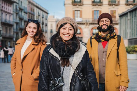 Foto de Grupo de amigos turísticos de pie en la calle de la ciudad europea sonriendo y mirando a la cámara. Retrato de tres personas viajeras felices turismo en un viaje de pie al aire libre. concepto de estilo de vida - Imagen libre de derechos