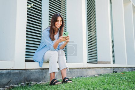 Téléchargez les photos : Joyeuse femme caucasienne souriant et naviguant sur Internet avec un téléphone mobile, textos e-mails, reliant les gens assis à l'extérieur. Jeune étudiante adolescente utilisant une application de téléphonie cellulaire sur le campus universitaire - en image libre de droit