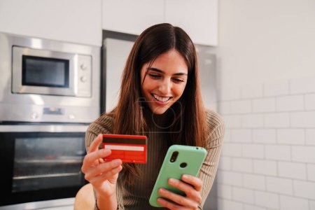 Photo for Young caucasian female with white perfect teeth smiling and using a cell phone app to purchase and pay with a credit card sitting at home kitchen. Woman spending money on online shopping on internet - Royalty Free Image