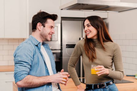 Téléchargez les photos : Jeune couple caucasien souriant boire du jus le matin debout à la maison cuisine. beau mari et femme brune riant et appréciant le petit déjeuner tenant un verre sain de boisson vitaminée - en image libre de droit