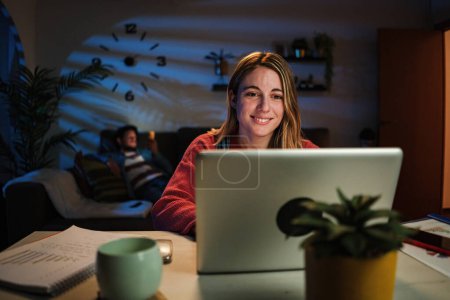 Téléchargez les photos : Jeune femme blonde caucasienne travaillant sur sa start-up avec ordinateur portable au bureau à la maison assis sur le bureau la nuit. Heureuse entrepreneure utilisant un ordinateur pour naviguer sur Internet avec connexion sans fil. Haut - en image libre de droit