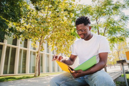 Photo for African american smart student reading a book sitting at university campus outdoors. Handsome concentrated young man with glasses studying and learning her notes of the lesson for high school exams - Royalty Free Image