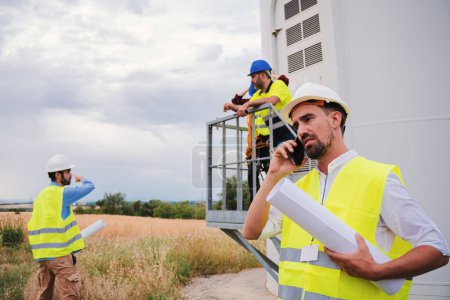 Photo for On foreground a caucasian engineer talkin by cell phone holding a blueprint, at background a group of technicians working. One industrial contractor having a smartphone call speaking about the project - Royalty Free Image