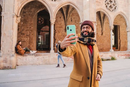 Téléchargez les photos : Heureux jeune homme adulte avec des lunettes et la barbe souriant et prendre un portrait selfie sur une ville européenne à partager sur les médias sociaux. Caucasien touriste mâle avoir plaisir à tirer une photo sur un voyage joutney - en image libre de droit