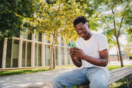 Téléchargez les photos : Heureux jeune homme afro-américain utilisant son téléphone portable à l'université, l'envoi de messages texte assis sur un banc à l'extérieur. Portrait d'un étudiant regardant l'écran de son téléphone portable sur le campus - en image libre de droit