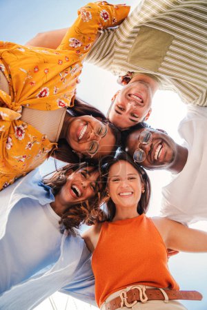 Foto de Vertical. Vista de bajo ángulo de un grupo de cinco adolescentes multirraciales sonriendo y mirando hacia abajo a la cámara al aire libre. Jóvenes estudiantes de diversas culturas étnicas abrazándose en un círculo - Imagen libre de derechos