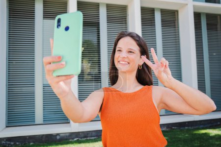 Foto de Mujer caucásica joven feliz sosteniendo el teléfono móvil tomando selfie usando el teléfono inteligente parado afuera en el campus de la universidad. Divertida estudiante disparando un autorretrato con un celular mostrando señal de paz - Imagen libre de derechos