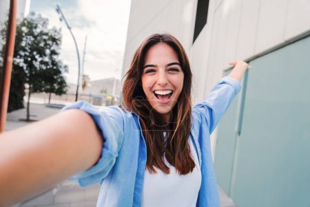 Téléchargez les photos : Joyeux jeune étudiante caucasienne regardant la caméra et prenant un portrait selfie s'amuser, debout à l'extérieur. Vue de face de la femme rieuse qui tourne une photo pour les médias sociaux sur le campus universitaire - en image libre de droit