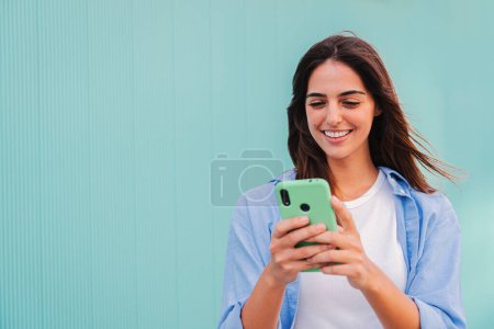 Téléchargez les photos : Souriant jeune femme heureuse en utilisant son smartphone et bavarder en ligne, ou en naviguant sur Internet à fond bleu sarcelle. Jeune étudiante caucasienne regarder des vidéos drôles avec téléphone mobile - en image libre de droit