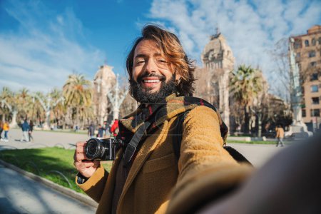 Foto de Joven bloguero de viajes divirtiéndose y riendo tomando un retrato de selfie y sonriendo en la ciudad de Europa sosteniendo una cámara en un viaje. Un hipster barbudo. Un turista en un escapo de fin de semana. Alto. - Imagen libre de derechos