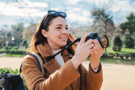 Téléchargez les photos : Jeune photographe souriante et photographiant une photo artistique lors d'une activité de week-end. Une femme hipster caucasienne prend plaisir à prendre une photo avec un appareil photo professionnel rétro lors d'un voyage de vacances - en image libre de droit