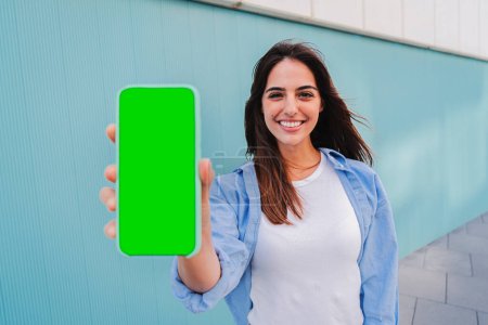 Photo for Isolated happy woman showing the green screen of her smartphone. Young smiling female holding a cellphone with chroma key. Mockup of caucasian joyful lady with mobile phone. Template for app or game - Royalty Free Image