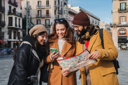 Photo for Group of tourist people sightseeing and searching a location on a map and on a smartphone app on a weekeand trip. Young travelers smiling looking together for directions on a guide visiting a city - Royalty Free Image