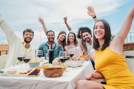 Foto de Grupo de amigos adultos jóvenes divirtiéndose y riéndose en la azotea de una cena. Gente multirracial feliz celebrando una reunión amistosa de almuerzo en casa bebiendo vino, sonriendo y comiendo juntos - Imagen libre de derechos