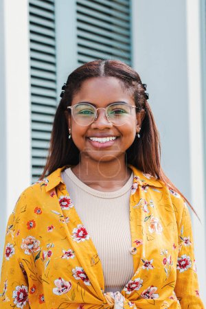 Photo for Vertical portrait of a hispanic happy teenage school girl with perfect white teeth smiling looking at camera santanding outdoors. Front view of young latin woman with eyeglasses laughing outside. High - Royalty Free Image