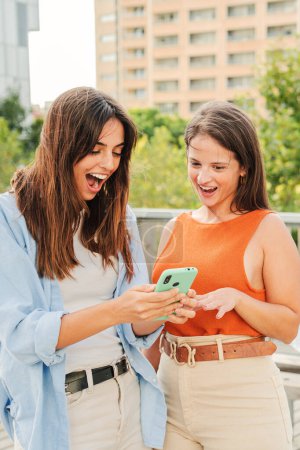 Foto de Vertical. Dos mujeres sonrientes alegres viendo y mostrando venta, noticias y redes sociales en su aplicación para teléfonos inteligentes fuera del campus universitario. Adolescentes jóvenes usando un teléfono celular juntas. Alto. - Imagen libre de derechos