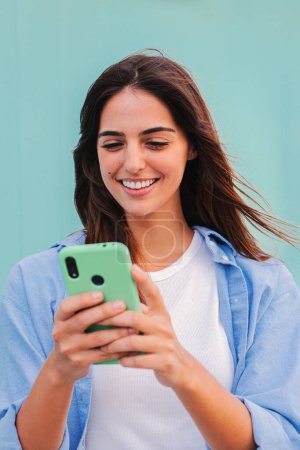 Téléchargez les photos : Portrait vertical d'une adolescente souriante et heureuse utilisant son smartphone et bavardant en ligne, ou naviguant sur Internet sur fond bleu sarcelle. Jeune étudiante dame regarder des vidéos drôles avec téléphone mobile - en image libre de droit