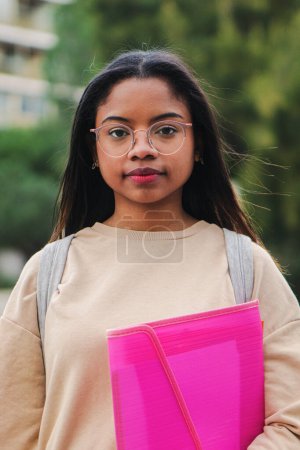 Photo for Vertical portrait of smart young latin school girl holding the notebook with pensive attitude. Serious hispanic teenage female standing outdoors at university capus looking at camera with his googles - Royalty Free Image