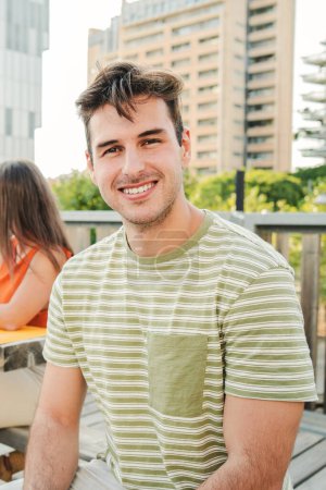 Téléchargez les photos : Portrait vertical d'un jeune étudiant positif et souriant regardant une caméra assise à l'extérieur du campus universitaire. Vue de face du bel homme riant avec une attitude joyeuse et amicale. Haut - en image libre de droit